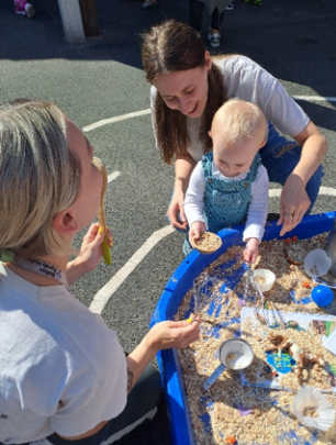 Image of a child, parent and a early years practitioner playing with the child together in a sandpit.